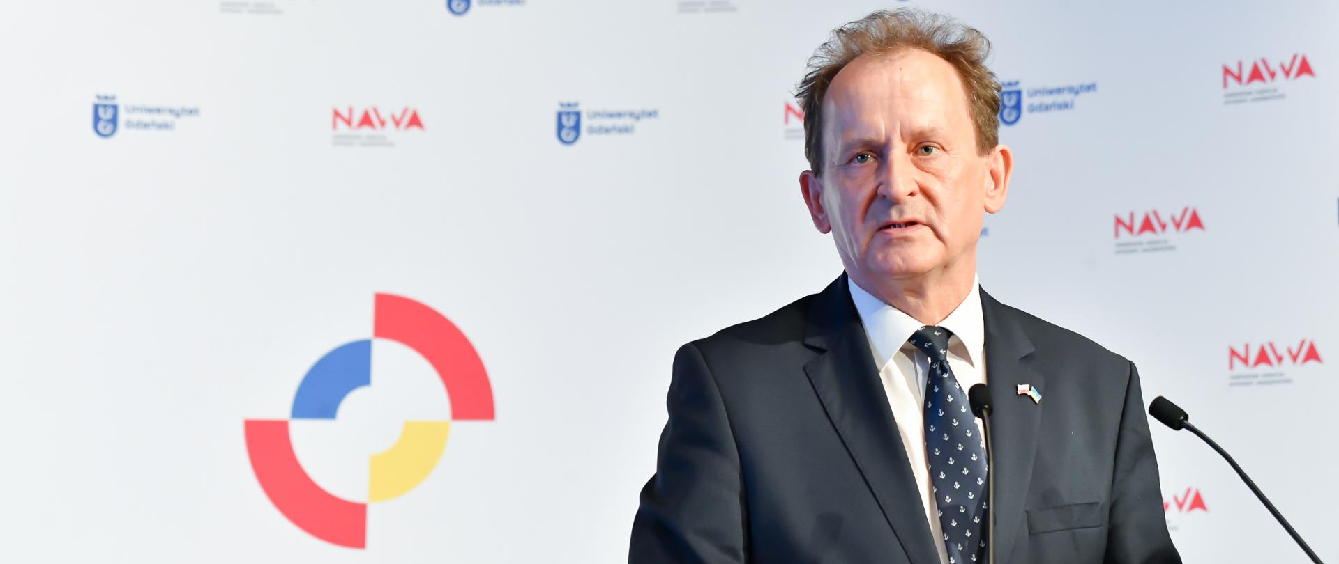 Wiceminister Włodzimierz Bernacki przemawia podczas konferencji. Stoi przy mównicy. Za sobą ma logo konferencji.