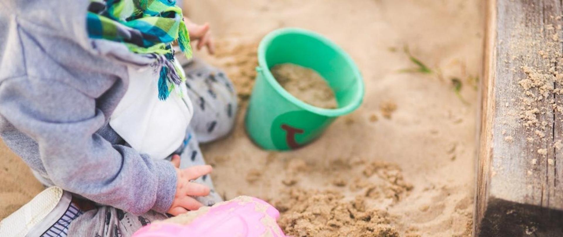 Na fotografii widzimy małe dziecko siedzące na piasku. Obok dziecka stoi zielone wiaderko z czerwonym obrazkiem na przedniej stronie, które jest częściowo wypełnione piaskiem. W tle rozpoznajemy drewnianą krawędź, być może jest to fragment piaskownicy. Zdjęcie skupia się na małym fragmencie przestrzeni, skupiając uwagę na czynnościach dziecka i teksturze piasku.