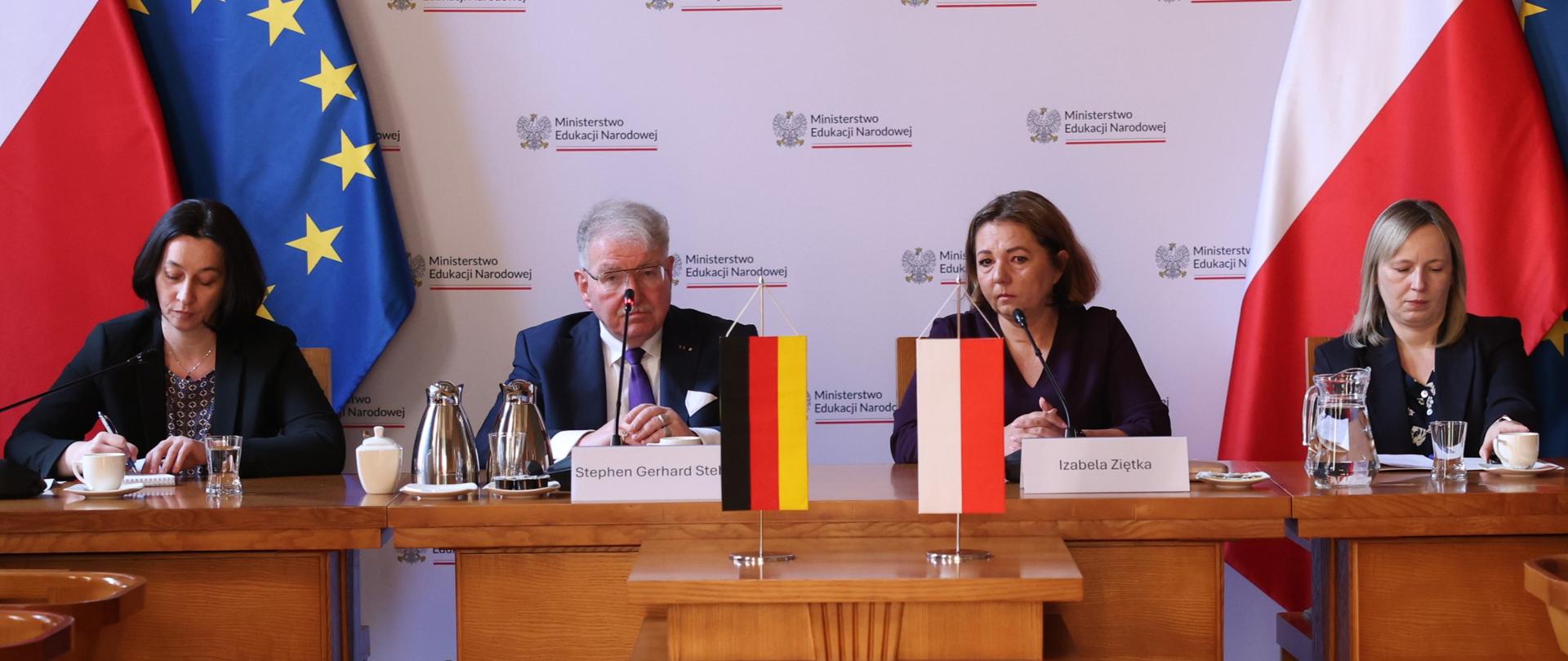 Oficjalne ujęcie czworga przedstawicieli delegacji siedzących przy stole prezydialnym. Przed nimi stoją tabliczki z nazwiskami: Stephen Gerhard Stehli oraz Izabela Ziętka. Na środku stołu umieszczono małe flagi Niemiec i Polski. Tło stanowi ścianka z powtarzającym się logotypem Ministerstwa Edukacji Narodowej oraz duże flagi Polski i UE.