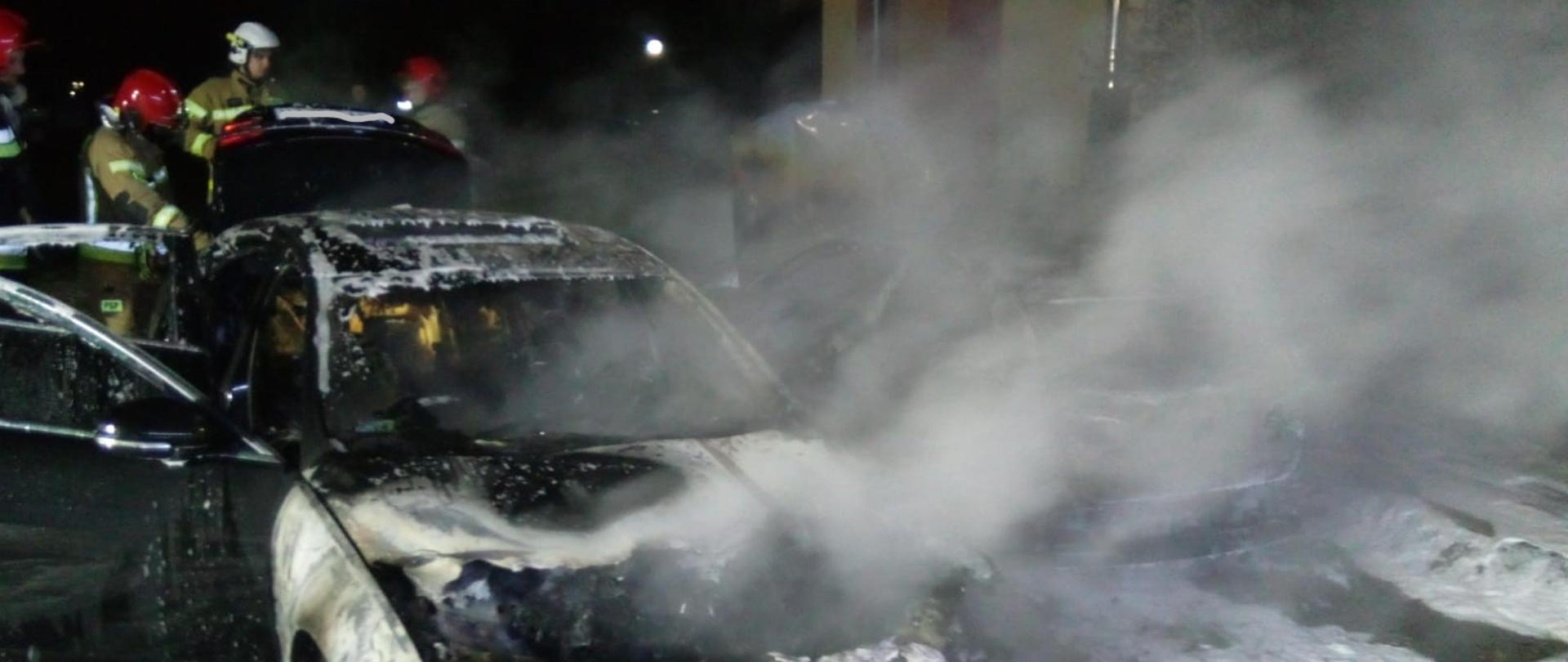 Zdjęcie przedstawia zniszczony w wyniku pożaru samochód z resztkami piany gaśniczej. Za autem pracuje grupa strażaków.