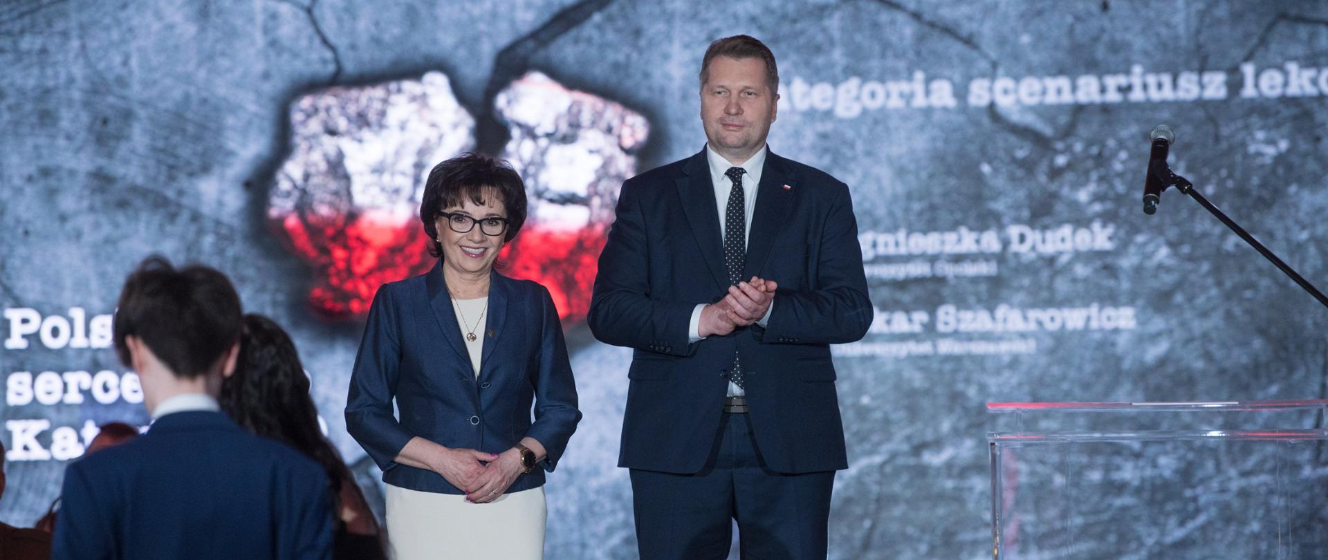 Minister Przemysław Czarnek wraz z Marszałek Sejmu Elżbietą Witek wręczają nagrody laureatom konkursu w kategorii scenariusz lekcji. Za nimi tło przedstawiające kamienną ścianę a na niej flagę Polski. Minister Przemysław Czarnek wręczył nagrody laureatom konkursu „Polskie Serce Pękło. Katyń 1940” 