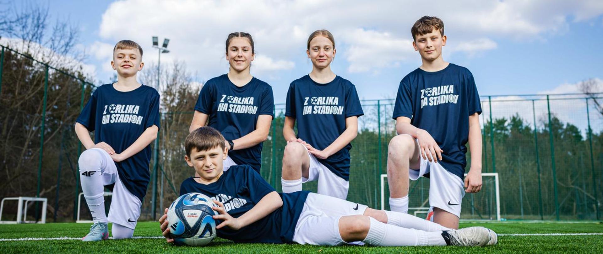 Group photo taken on a football pitch: two boys and two girls wearing navy blue "From Orlik pitch to the Stadium" t-shirts and white shorts kneel on one knee. A third boy lies on his side on the grass in front of them holding a football.