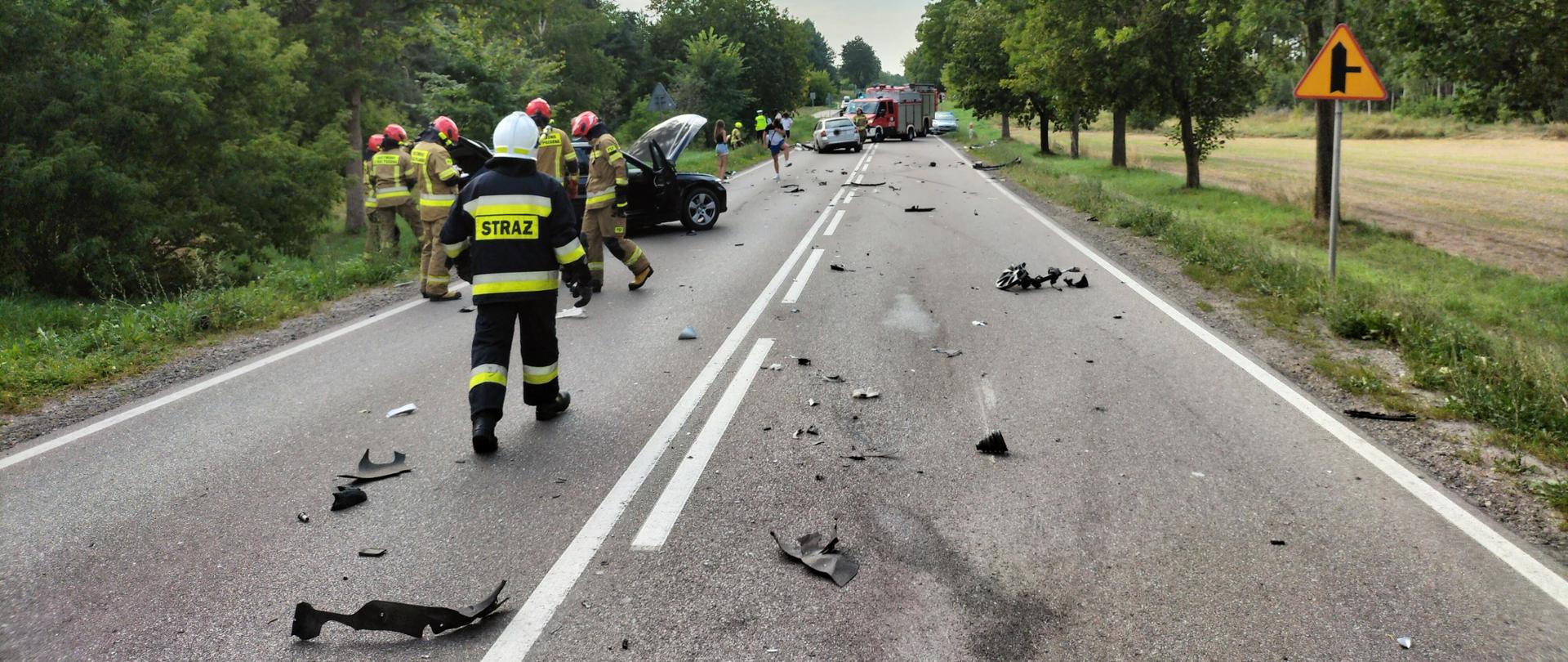 Strażacy stoją przy rozbitym samochodzie. W tle inny rozbity pojazd oraz samochody straży pożarnej.