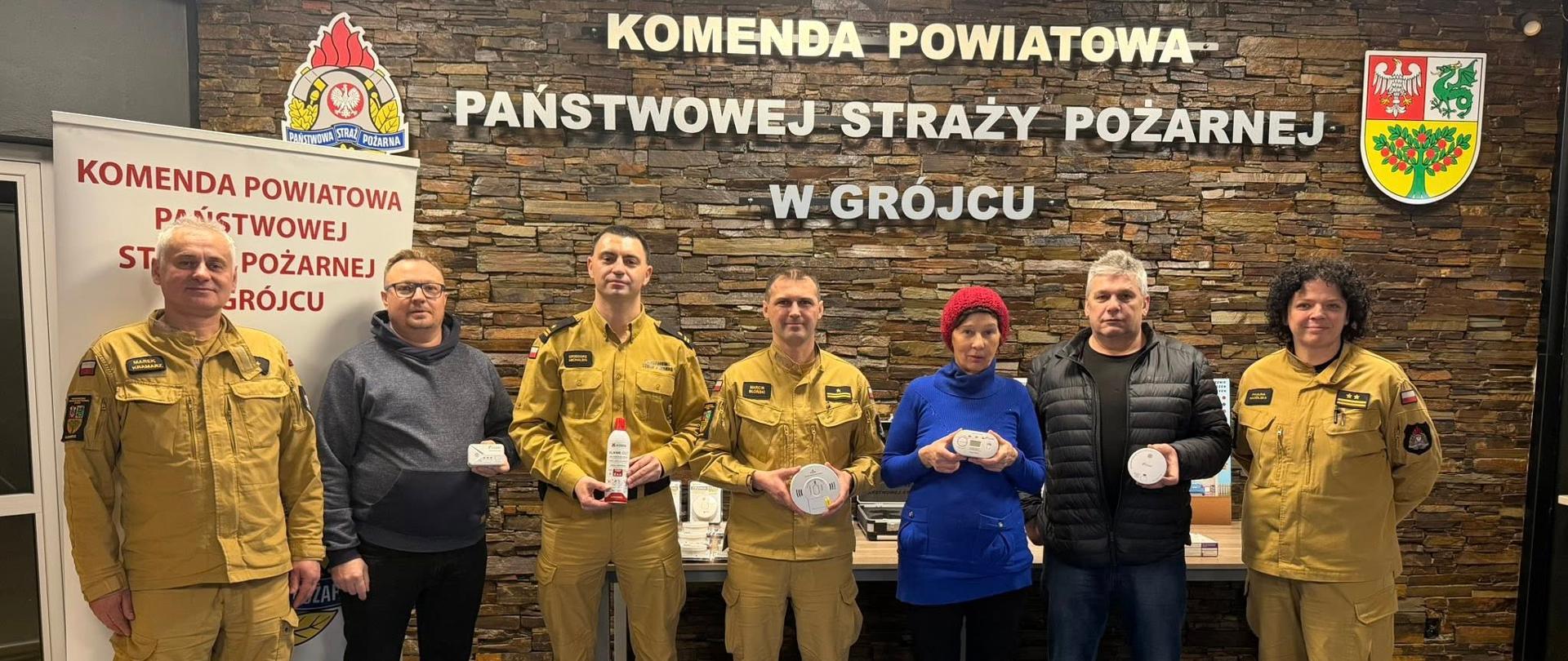 Na zdjęciu grupowym stoi osiem osób ustawionych w jednym rzędzie.
Część osób ma na sobie żółte mundury strażackie, a część ubrania cywilne (kurtki, swetry).
Kilka osób trzyma w rękach okrągłe czujniki dymu lub czadu oraz jedno podłużne urządzenie.
Wszyscy stoją przodem do aparatu. Za nimi znajduje się ta sama kamienna ściana z napisem
„Komenda Powiatowa Państwowej Straży Pożarnej w Grójcu”.
Zdjęcie ma charakter oficjalny i dokumentacyjny.