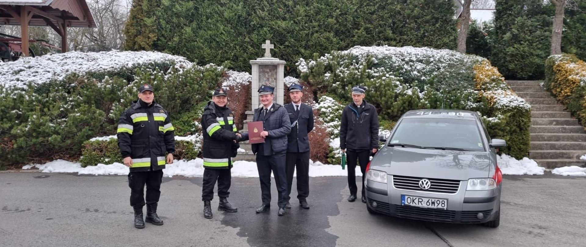 1. Tło stanowi porośnięta krzewami skarpa, częściowo ośnieżona. W środku skarpy kapliczka ze św. Florianem. Na tle skarpy po prawej ustawiony frontem samochód operacyjny VW Passat w kolorze ciemnoszarym. W centrum zdjęcia ustawieni od lewej dwaj strażacy PSP w ubraniach dowódczo-sztabowych. Dalej trzej członkowie OSP Rozwadza w mundurach wyjściowych i kurtkach. Komendant Powiatowy PSP podaje prawą rękę prezesowi OSP. Prezes OSP w lewej ręce trzyma okolicznościową teczkę z aktem przekazania. 