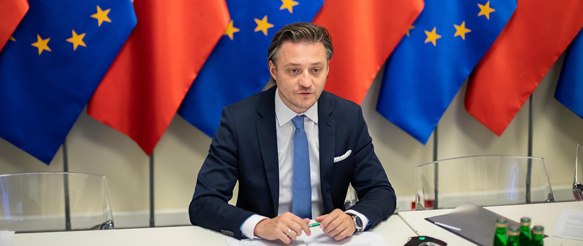 Na zdjęciu widać wiceministra Bartosza Grodeckiego siedzącego za stołem w trakcie wideokonferencji. W tle widać rząd flag Polski i UE.