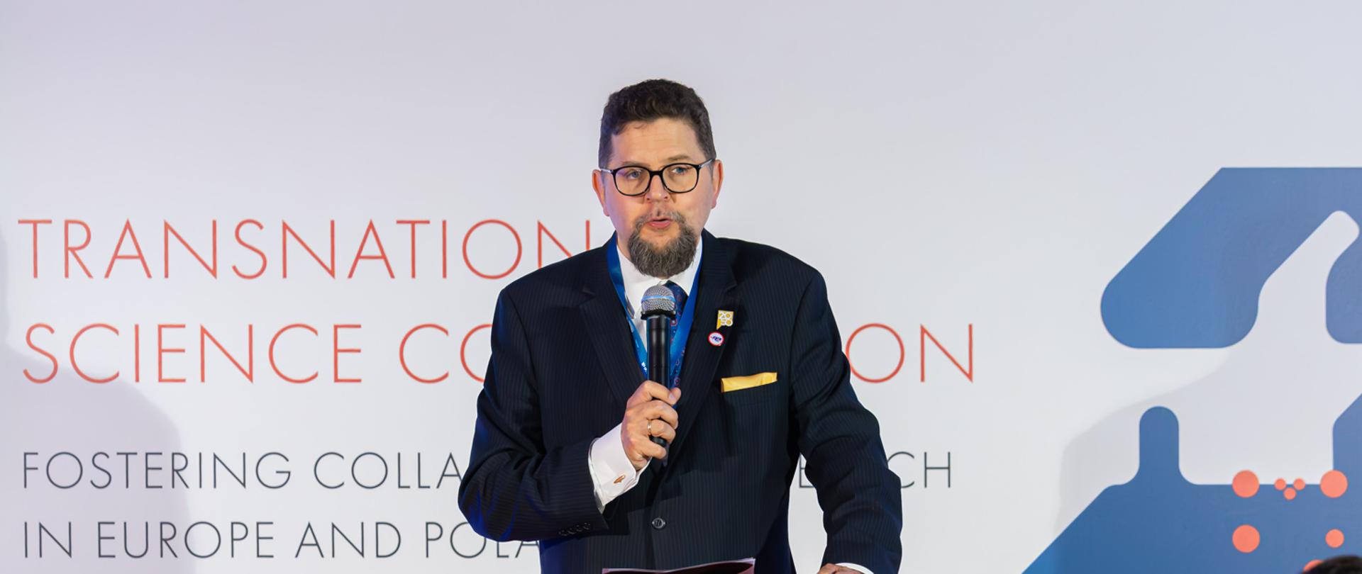 Wiceminister Szeptycki stoi za mównicą i mówi do trzymanego w ręku mikrofonu.