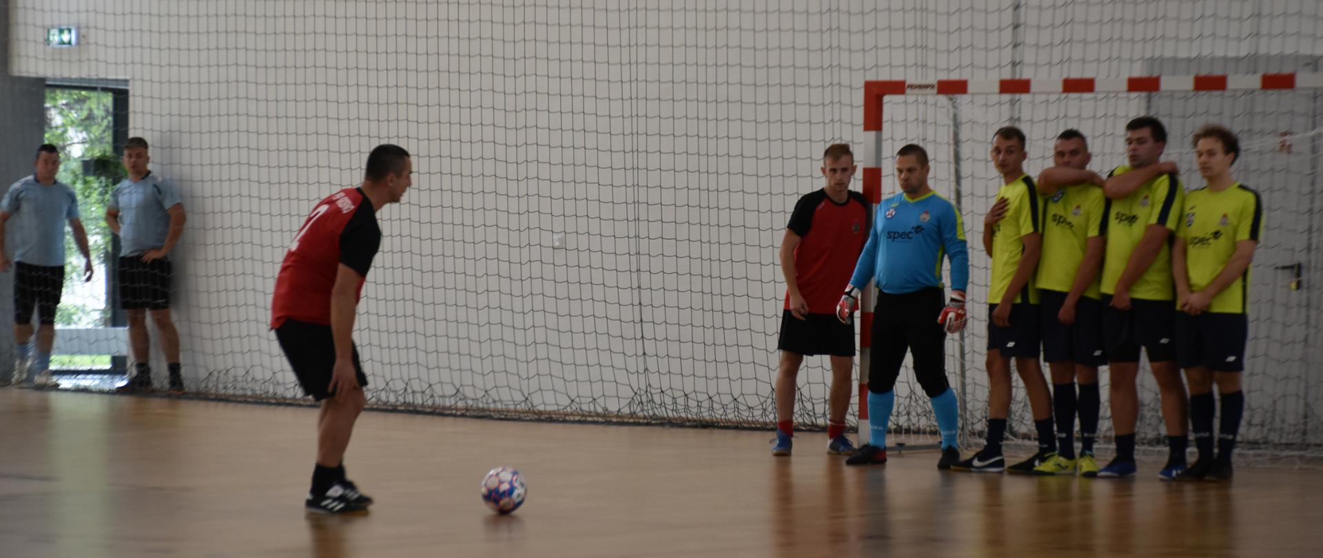 Zdjęcie przedstawia wszystkie wnętrze hali sportowej zawodnik w stroju koloru czerwonego wykonuje pośredni rzut wolny. zawodnicy w strojach seledynowych ustawieni na linii bramkowej