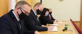 Na zdjęciu cztery osoby, zasiadające za stołem - podpisują dokumenty. Kadr boczny.