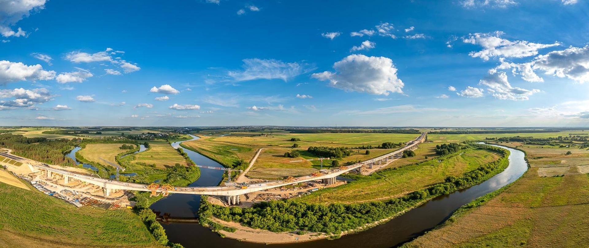 Widok z lotu ptaka na wielką plac budowy mostu nad meandrującą rzeką. Rzeka wygina się przez krajobraz, zielone pola i skupiska drzew po obu stronach. Na placu budowy widać wiele żurawi, kilka częściowo ukończonych odcinków mostu z widocznymi rusztowaniami oraz liczne pojazdy budowlane. Niebo jest bezchmurne, co sugeruje dobre warunki pogodowe do prac budowlanych.