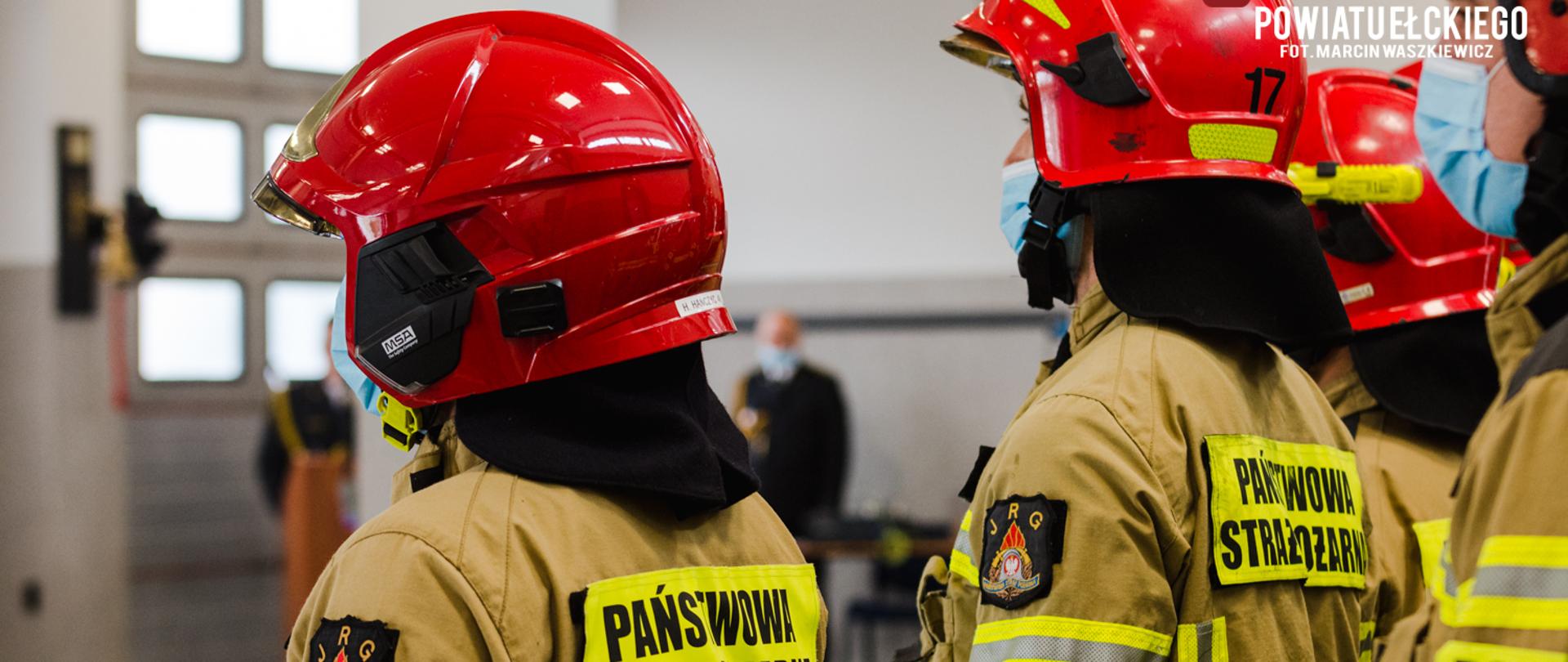 Zdjęcie przedstawia strażaków w szyku stojących tyłem do fotografa