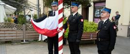 Widok z przodu. Trzech strażaków PSP tworzących poczet flagowy w trakcie podnoszenia flagi państwowej na maszt.