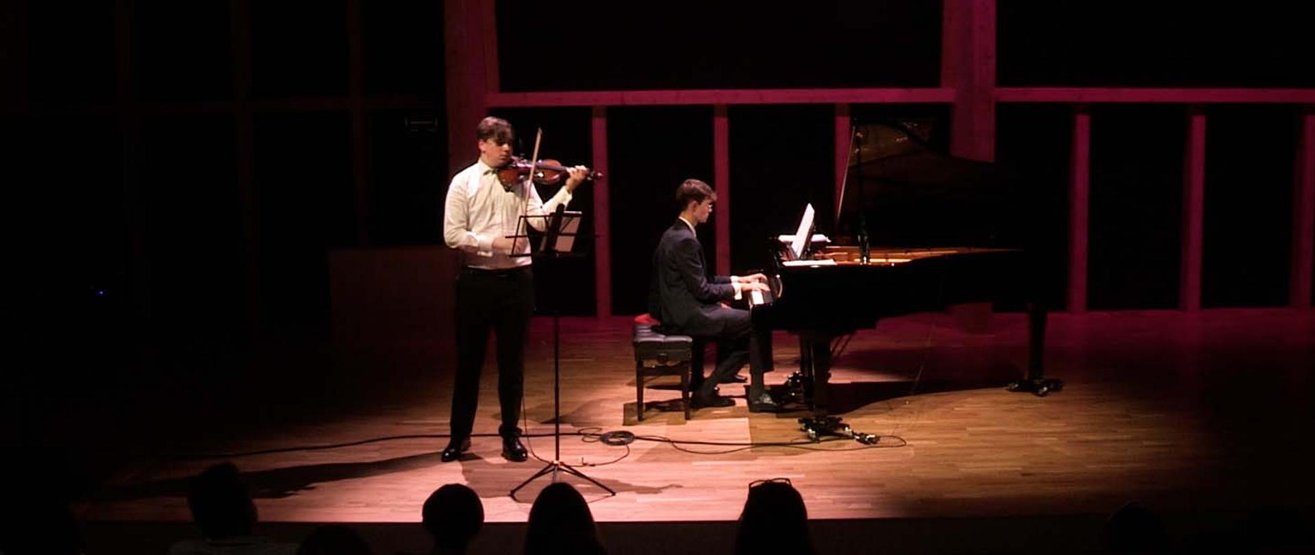 Dwaj mężczyźni występujący na scenie, jeden stoi z lewej strony i gra na skrzypcach, drugi z tyłu gra na fortepianie.