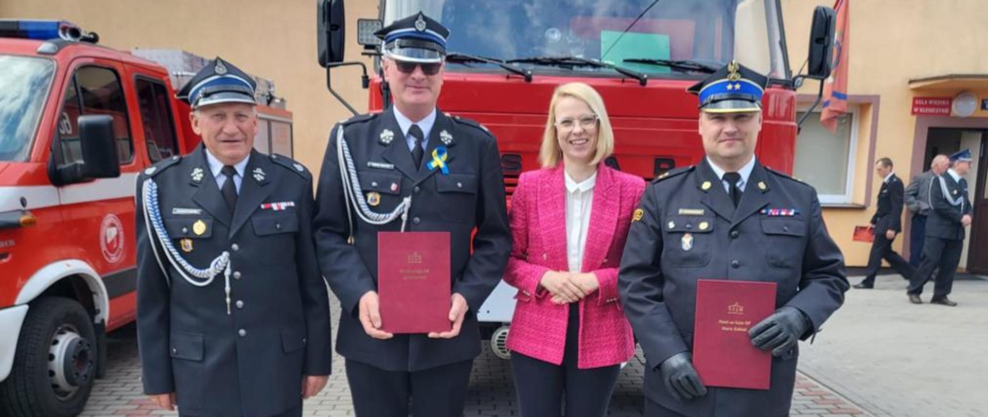 Zdjęcie przedstawia poseł Martę Kubiak komendanta powiatowego Prezesa oraz jednego z druhów na tle samochodu strażackiego