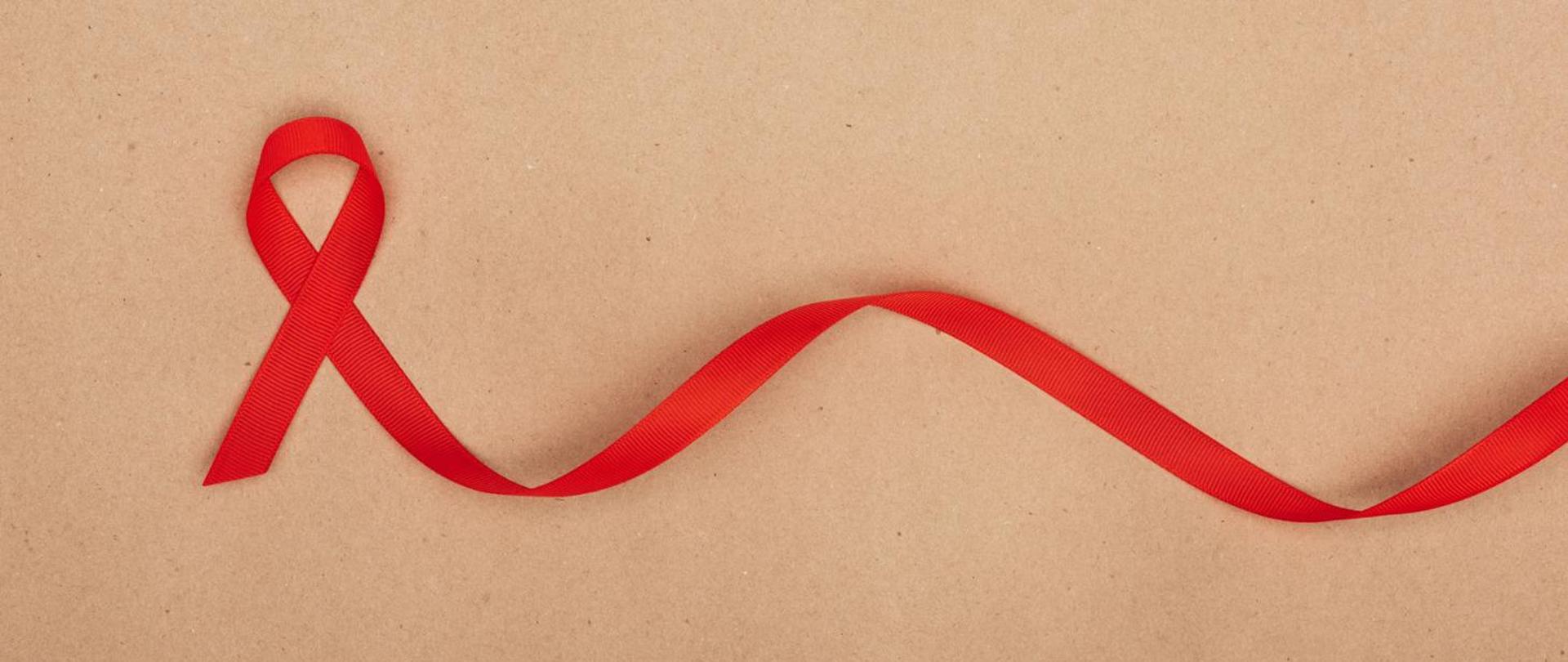 Na zdjęciu widoczna jest czerwona wstążka, która jest symbolem świadomości na temat HIV i AIDS. Wstążka jest umieszczona na tle jednolitego brązowego papieru. Ma klasyczny kształt litery "V" i rozciąga się na płaskiej powierzchni, tworząc subtelną krzywą. Czerwona wstążka jest powszechnie używana do podnoszenia świadomości na temat zapobiegania HIV oraz wsparcia dla osób żyjących z tą chorobą.