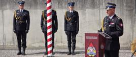 Komendant Wojewódzki przemawia z mównicy ustawionej na placu wewnętrznym komendy do zebranych strażaków. W tle poczet flagowy stojący w mundurach galowych przy maszcie. 