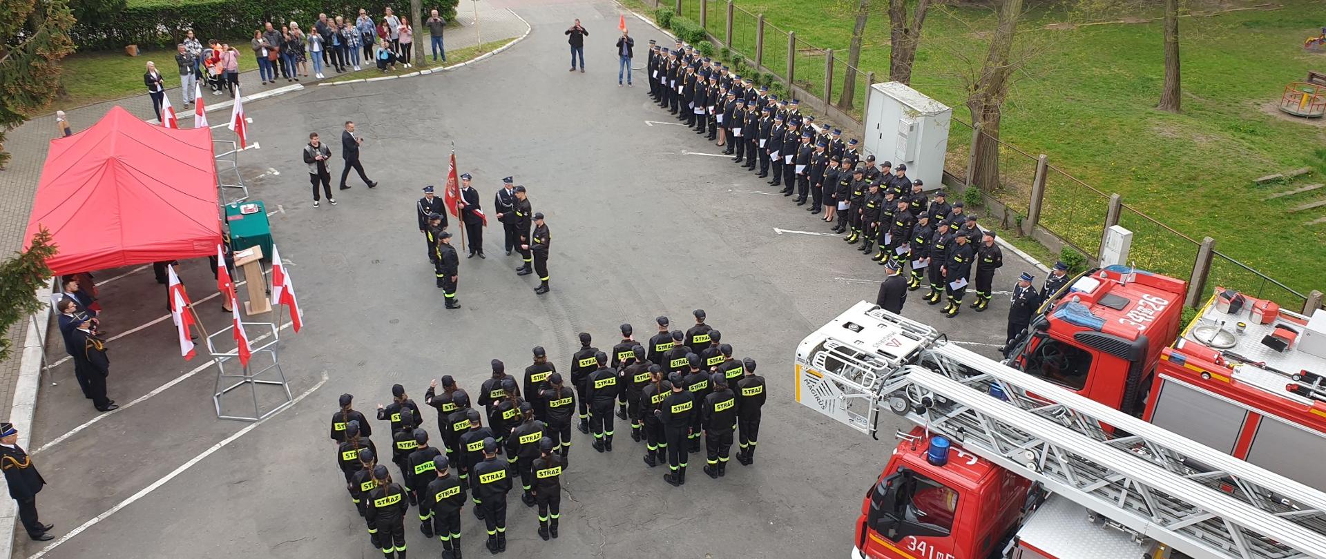 Widok na plac komendy z góry. Strażacy stoją w dwuszeregu. po prawej stronie widoczne pojazdy pożarnicze, po lewej stronie czerwony namiot pod którym znajdują się zgromadzeni goście. 