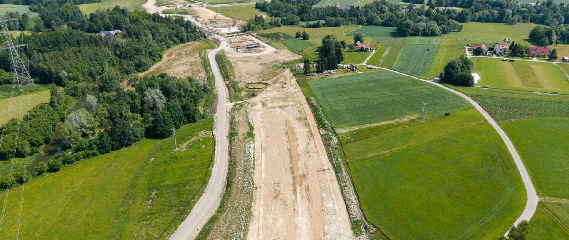 Widok z lotu ptaka na budowę drogi ekspresowej prowadzoną wśród zielonych pól i zadrzewień. W centrum zdjęcia widoczny jest szeroki pas przygotowanego terenu pod przyszłą jezdnię z wykonanymi robotami ziemnymi. Po lewej stronie biegnie tymczasowa droga technologiczna, a obok niej znajdują się gęste skupiska drzew oraz niewielki ciek wodny. W górnej części fotografii widoczny jest obiekt inżynierski w budowie oraz zaplecze robót. Po prawej stronie znajdują się pola uprawne, lokalne drogi i pojedyncze zabudowania mieszkalne. Zdjęcie wykonano w słoneczny dzień, ukazując postęp prac drogowych na tle pagórkowatego krajobrazu.