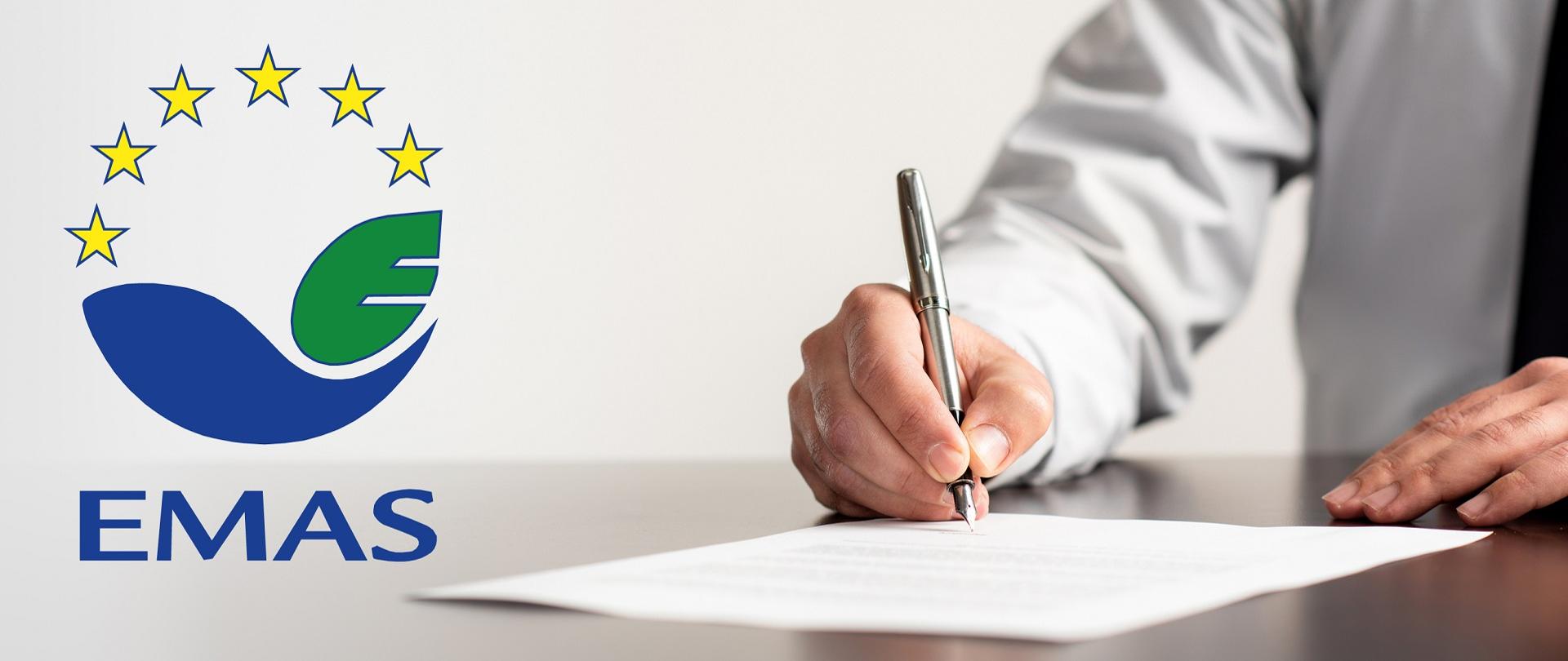 Zbliżenie na dłoń mężczyzny, który piórem podpisuje dokument. Z lewej strony logotyp EMAS.