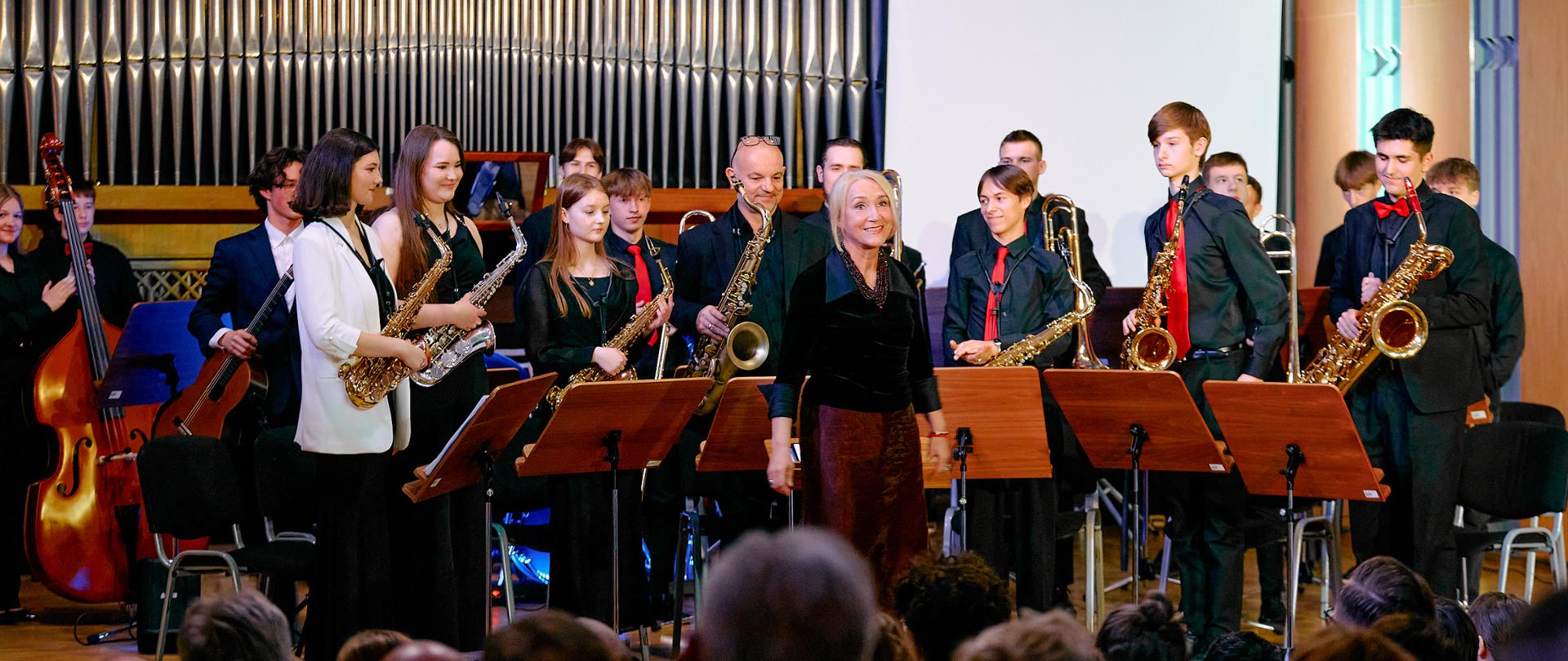 Na zdjęciu widzimy moment zakończenia koncertu lub występu zespołu muzycznego j big bandu – w sali koncertowej. Na pierwszym planie znajduje się grupa widzów siedzących tyłem do obiektywu. Kilka osób klaszcze. Na scenie, przed dużymi organami piszczałkowymi, stoi około 15 młodych muzyków – chłopców i dziewcząt – w eleganckich, koncertowych strojach. Większość ubrana jest na czarno z czerwonymi akcentami, takimi jak krawaty czy muszki. Instrumenty: muzycy trzymają saksofony, puzon, gitarę i kontrabas. Przed nimi stoją pulpity nutowe. Na środku sceny, przed muzykami, stoi kobieta – prawdopodobnie dyrygentka lub prowadząca koncert. Uśmiecha się do publiczności. W tle, za muzykami, znajduje się ekran projekcyjny i monumentalne piszczałki organowe, które dodają sali klasycznego, dostojnego charakteru. Zdjęcie oddaje ciepłą, podniosłą atmosferę zakończenia koncertu – moment, w którym zespół otrzymuje brawa za swój występ.