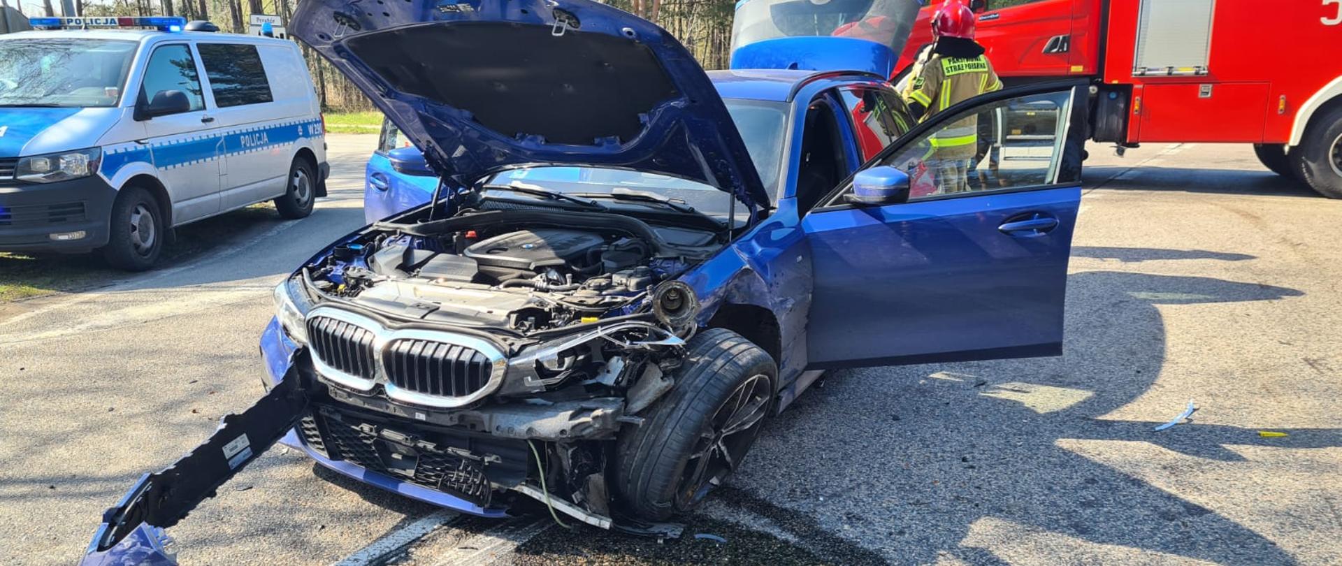 Zdjęcie przedstawia uszkodzony samochód osobowy marki BMW koloru granatowego. samochód stoi na środku drogi z otwartą maską. W tle widać samochód pożarniczy oraz radiowóz Policji.