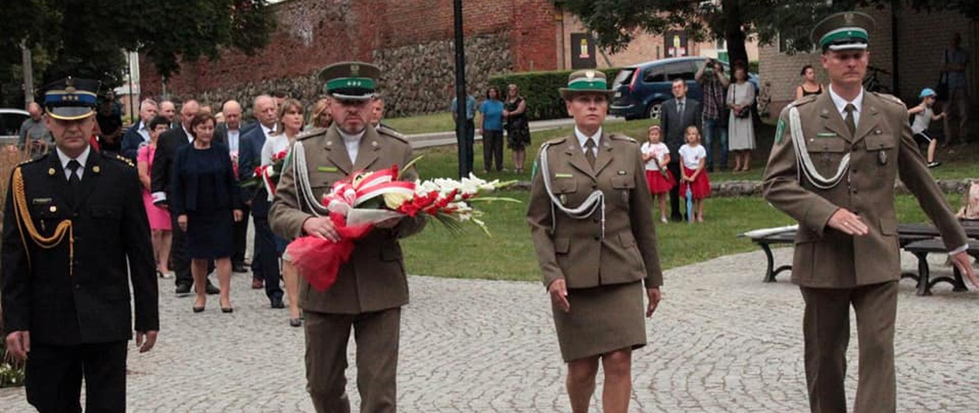 Zdjęcie zrobione przed przed pomnikiem na placu. W szeregu idą funkcjonariusze od lewej: komendant Powiatowy PSP w Kętrzynie w czarnym mundurze galowym z rogatywką na głowie, drugi i czwarty funkcjonariusz Straży Granicznej w zielonym mundurze galowym z rogatywką na głowie (spódnica zamiast spodni) oraz trzecia w kolejności w szeregu funkcjonariuszka straży granicznej w zielonym mundurze galowym z rogatywką na głowie. Drugi funkcjonariusz trzyma w rękach kwiaty. W tle inni uczestniczy uroczystości.