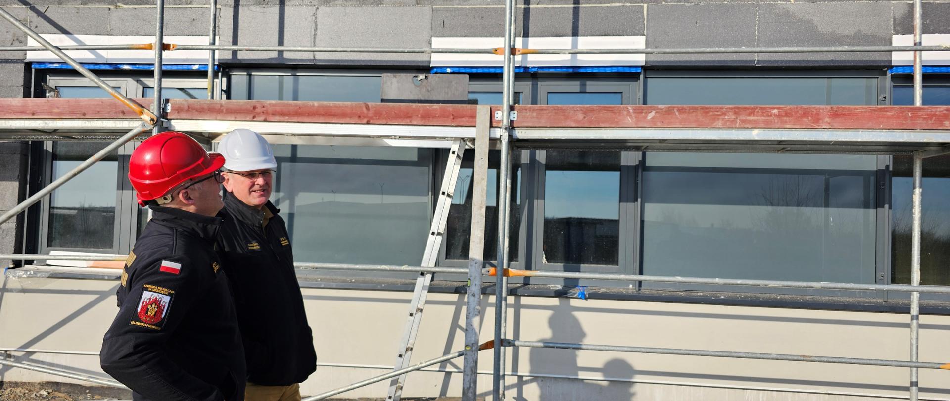 Dwaj strażacy w piaskowo-czarnych mundurach i kaskach na głowie stojący i rozmawiający przy ścianie z oknami budowanego obiektu.