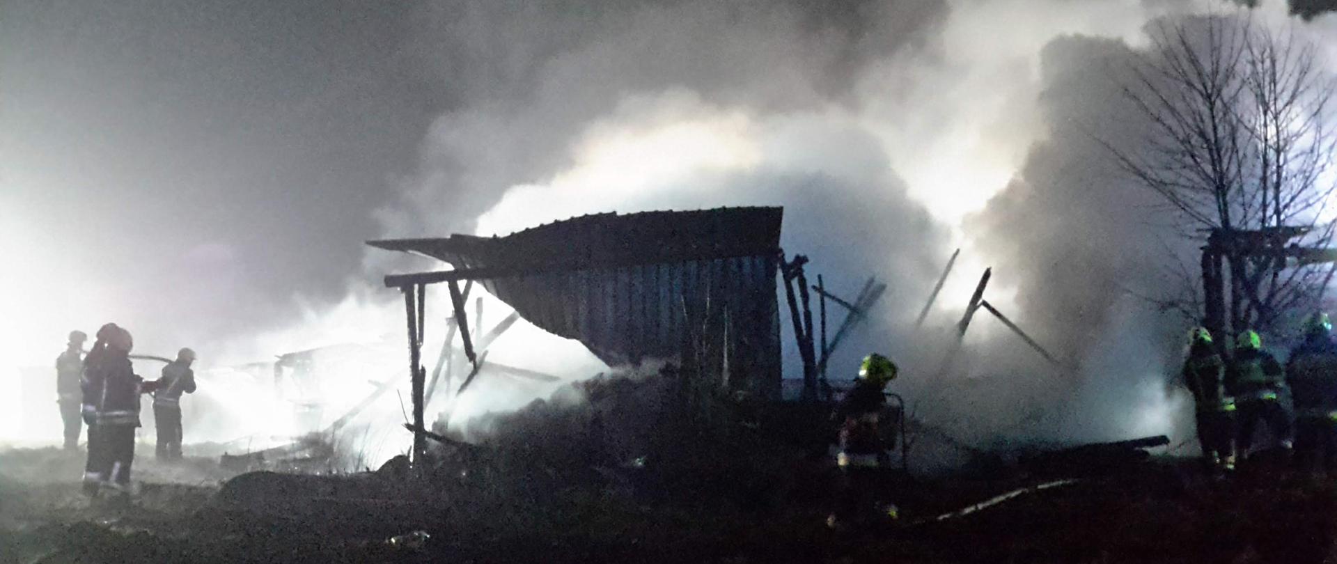 Pora nocna, dwóch strażaków trzymając wąż strażacki gasi pożar. W tle drewniane spalone elementy stodoły i unoszące się kłęby dymu.