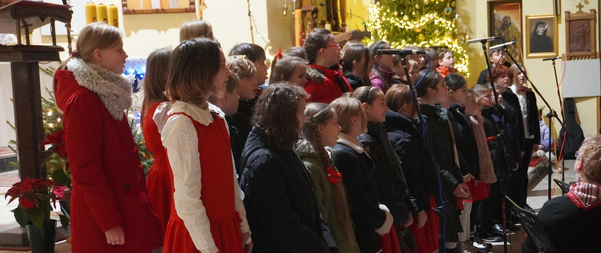 Szkolny chór podczas występu na koncercie kolęd w Redzie. Dzieci stoją przed mikrofonami i śpiewają. Większość w zimowych kurtkach z uwagi na niską temperaturę w kościele.