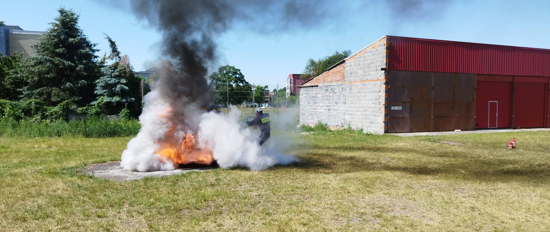 Na zdjęciu widać ogień, dym oraz strażaka. Zdjęcie wykonane w ciągu dnia. Słonecznie. Niebo błękitne. W tle zabudowania.