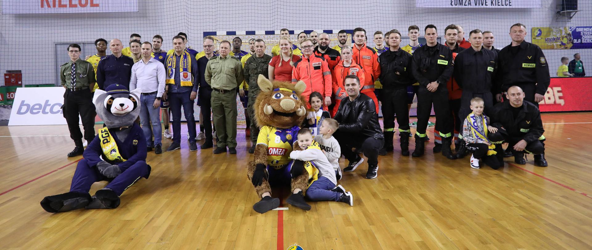 Na zdjęciu widzimy grupę kibiców przedstawicieli służb mundurowych z zawodnikami zespołu Łomża Vive Kielce na boisku. Część kibiców jest w mundurach różnych formacji - Państwowej Straży Pożarnej, Policji, Straży miejskiej, Straży Granicznej, Zespołu Ratownictwa Medycznego.