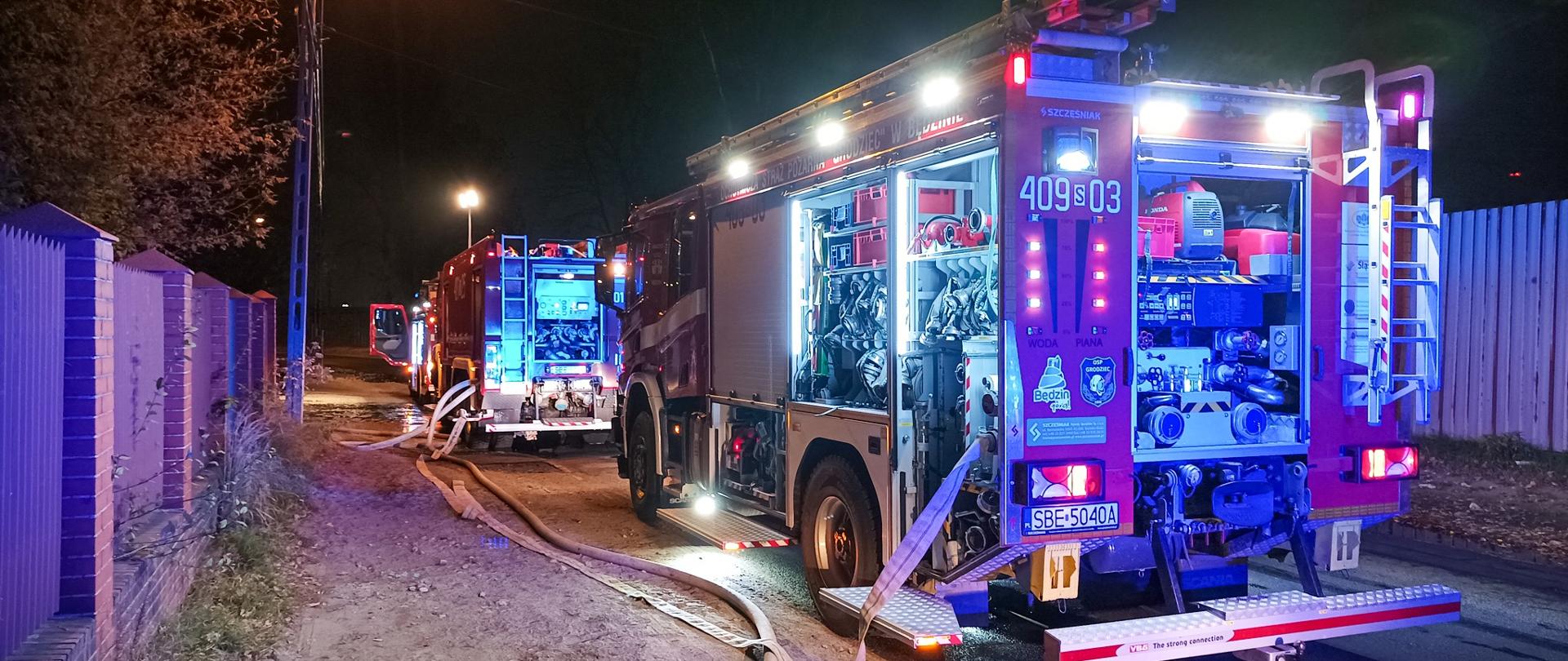 Zdjęcie przedstawia trzy pojazdy gaśnicze ustawione na drodze dojazdowej do posesji przy której miało miejsce zdarzenie. Przy samochodach widać połączone linie wężowe którymi podawano wodę do miejsca pożaru. 