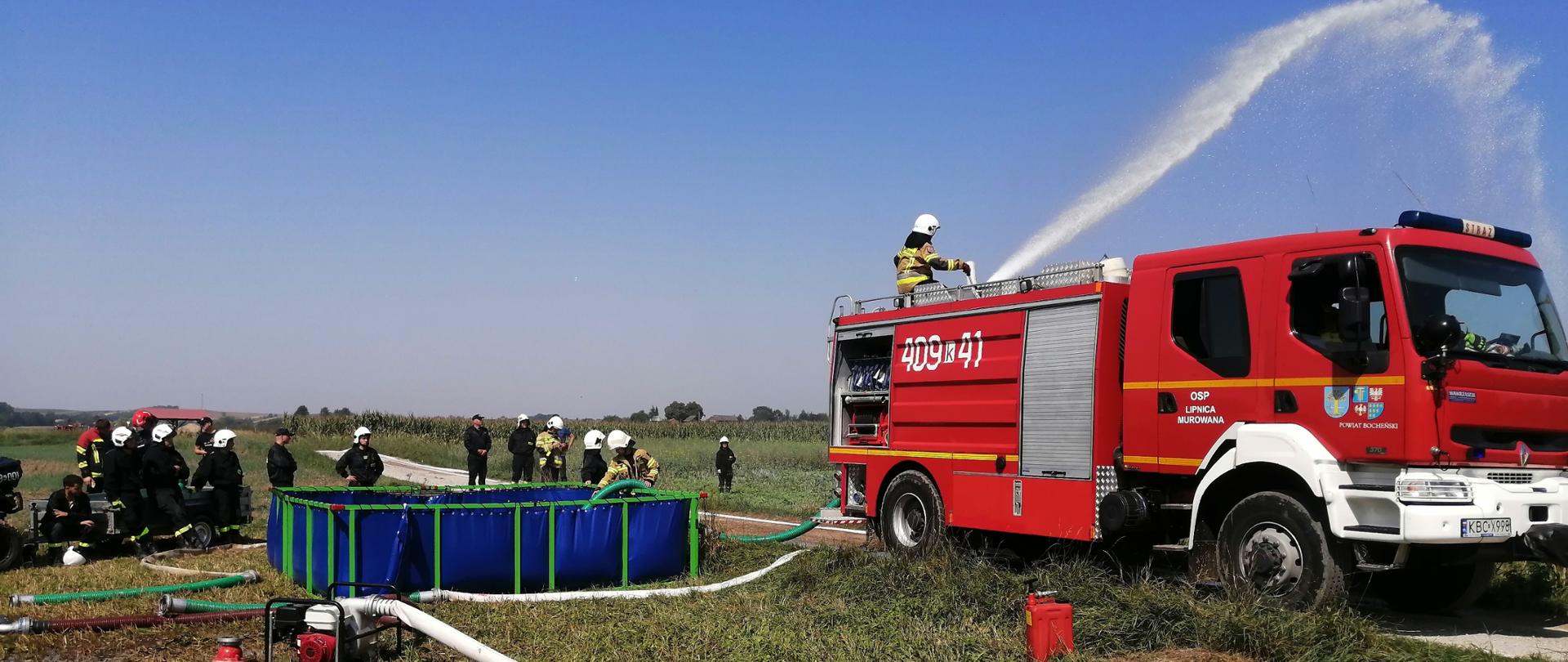 Zdjęcie na pierwszym planie ukazuje wóz strażacki stojący obok zbiornika na wodę. Jeden ze strażaków stoi na dachu wozu i kieruje prądem wody z działka wodnego zamocowanego na dachu samochodu. W tle widać strażaków ,którzy przyglądają się ćwiczeniom. Niebo jest bezchmurne