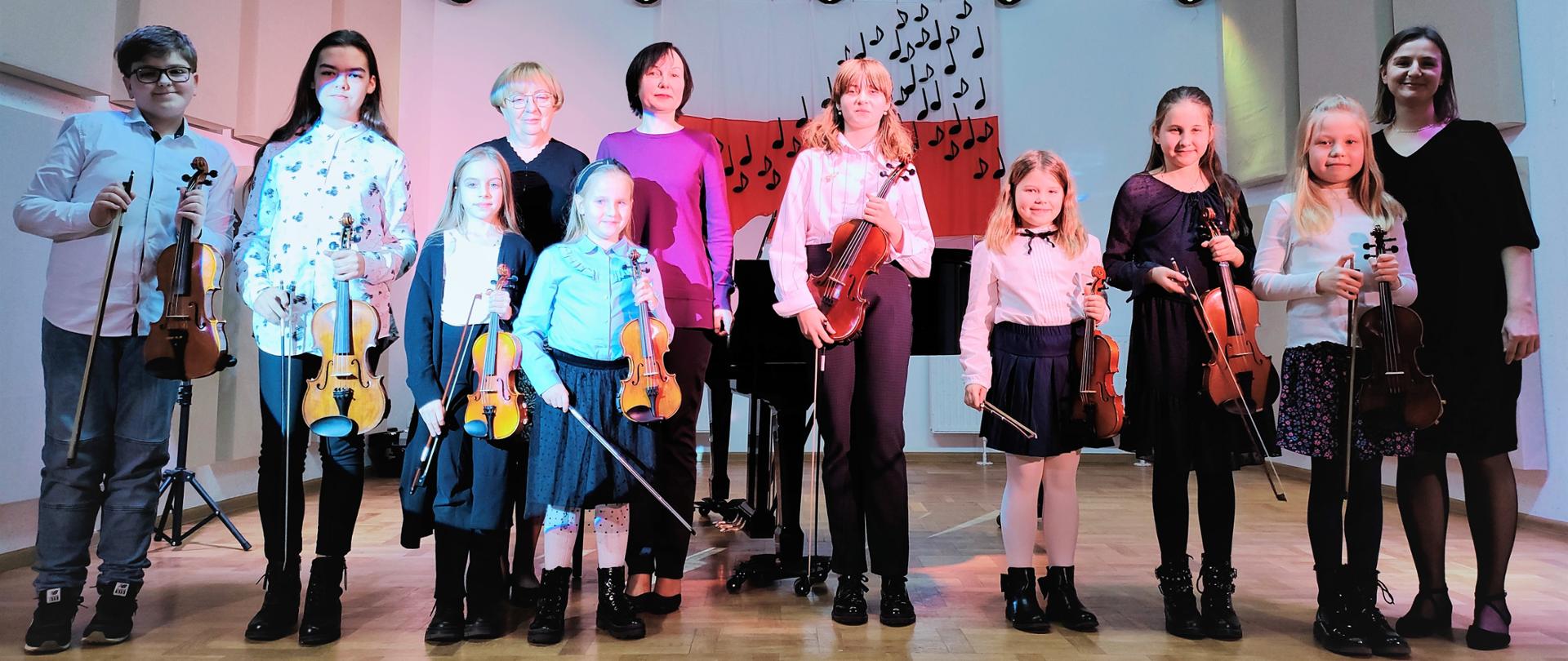 Zdjęcie przedstawia uczniów klasy skrzypiec stojących na scenie szkoły. Dzieci trzymaja w rękach skrzypce. Wraz z nimi stoją nauczyciele. Za uczniami na środku sceny stoi czarny fortepian, a na tylnej ścianie wisi flaga Polski z czarnymi nutami.