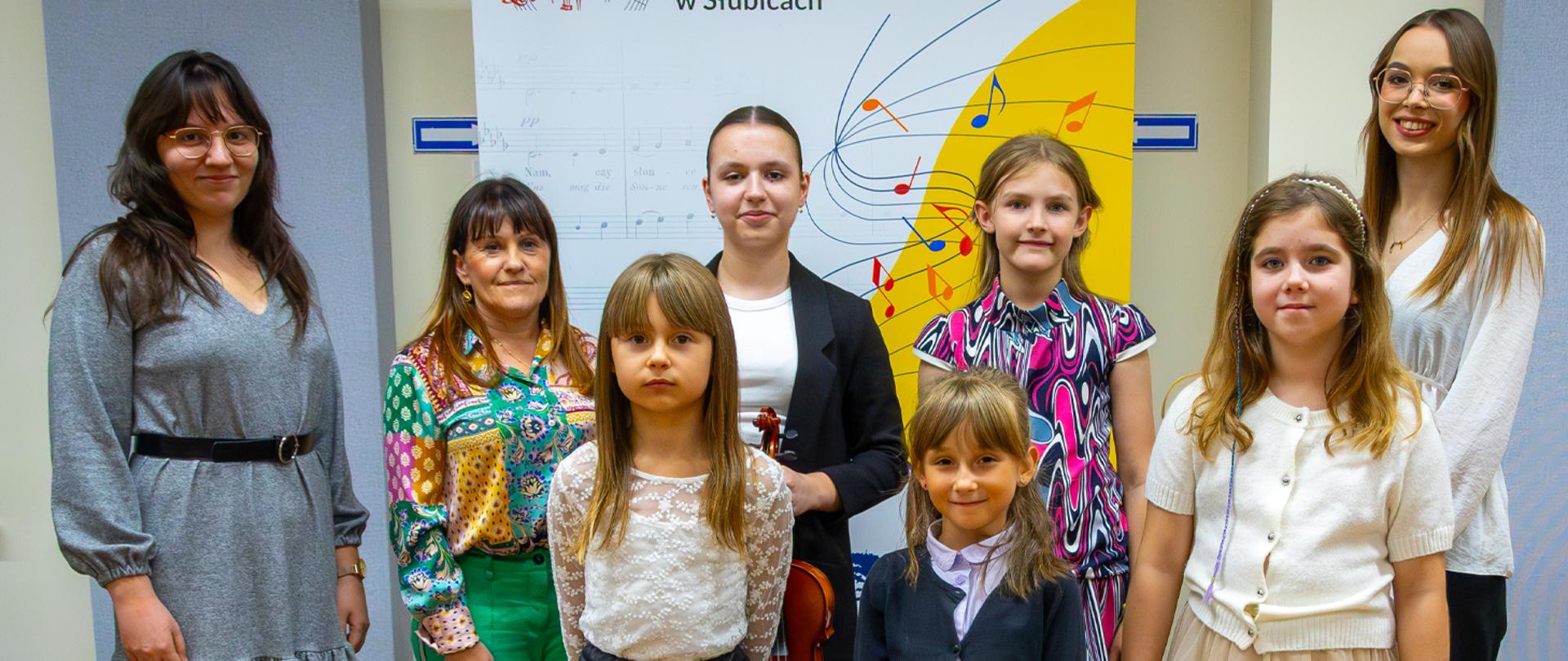 Cztery uczennice i trzy nauczycielki stoją w auli szkolnej na tle baneru szkolnego.