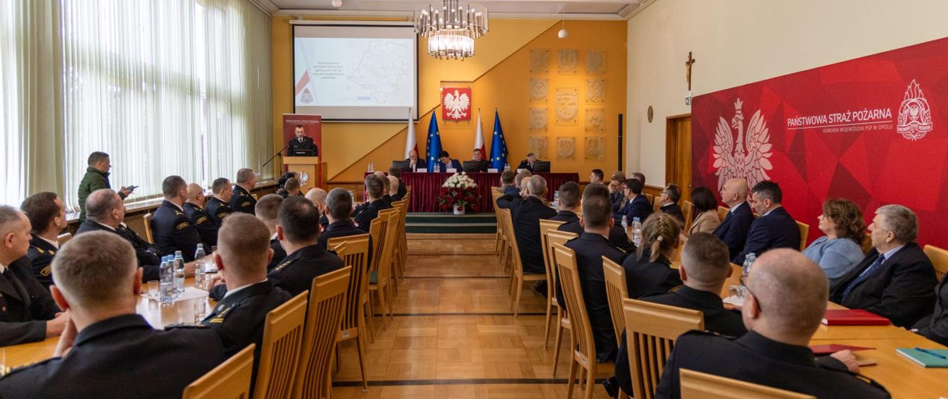 Zastępca Komendanta stoi na mównicy i przemawia do mikrofonu. Przed mównicą przy stołach siedzą funkcjonariusze PSP oraz zaproszeni goście.