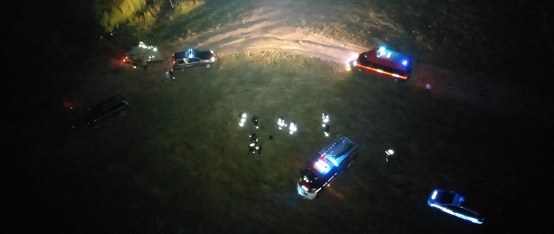 Zdjęcie robione z drona. Ujęcie z lotu ptaka. Na fotografii zrobionej w ciemnościach widać krąg światła rzucany przez zamontowane na dronie oświetlenie a w nim cztery samochody z błyskającymi niebieskimi światłami. Trzy z nich do pojazdy straży pożarnej , do jednego doczepiona jest przyczepka z łodzią. Czwarty samochód do radiowóz policji. Na zdjęciu widoczny jest jeszcze jeden pojazd bez oznaczeń, z wyłączonymi światłami. Pomiędzy pojazdami widoczne są osoby ubraniach z odblaskowymi elementami. Dwie osoby są ubrane w cywilne rzeczy. Łącznie jest trzynaście osób. Teren akcji jest pokryty trawą i widoczny jest częściowo element piaszczystej drogi.
