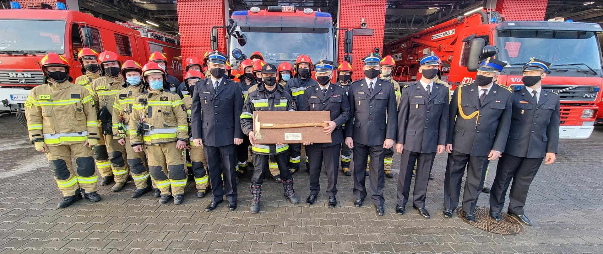 26.02.2021 Ostatnia zmiana służby asp. sztab. Tomasz Kołodziński. Na zdjęciu w tel samochody pożarnicze w garażu. Na pierwszym planie strażacy w dwuszeregu wszyscy w mundurach wyjściowych i bojowych. Na środku Tomasz Kołodziński Z-ca dowódcy JRG Legionowo.