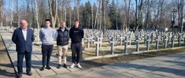 Trzej uczniowie i nauczyciel stoją na cmentarzu, w tle liczne krzyże nagrobne.