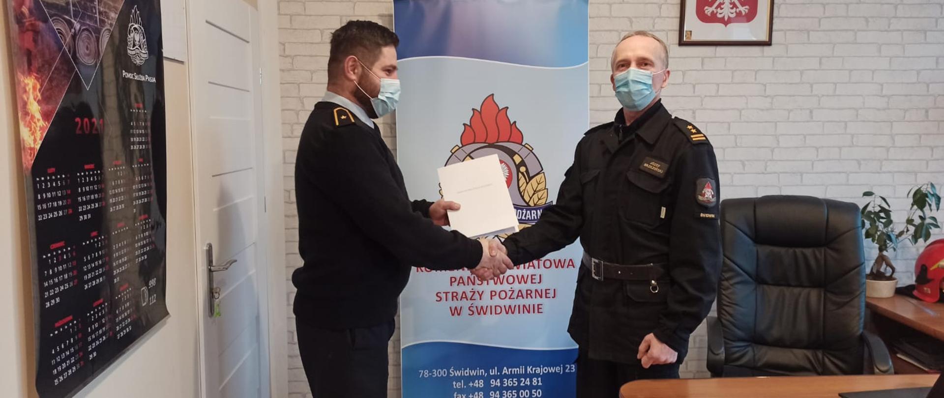 Zdjęcie przedstawia dwóch mężczyzn ubranych w mundury służbowe oraz maseczki chirurgiczne, jeden z nich trzyma w rękach dokumenty. Za nimi widnieje baner z logiem PSP oraz napisem: "Komenda Powiatowa Państwowej Straży Pożarnej w Świdwinie". Za banerem widzimy ścianę z tapetą w szare cegły. Na ścianie wisi Godło Rzeczypospolitej Polskiej. Zdjęcie wykonano w gabinecie Komendanta Powiatowego PSP w Świdwinie.