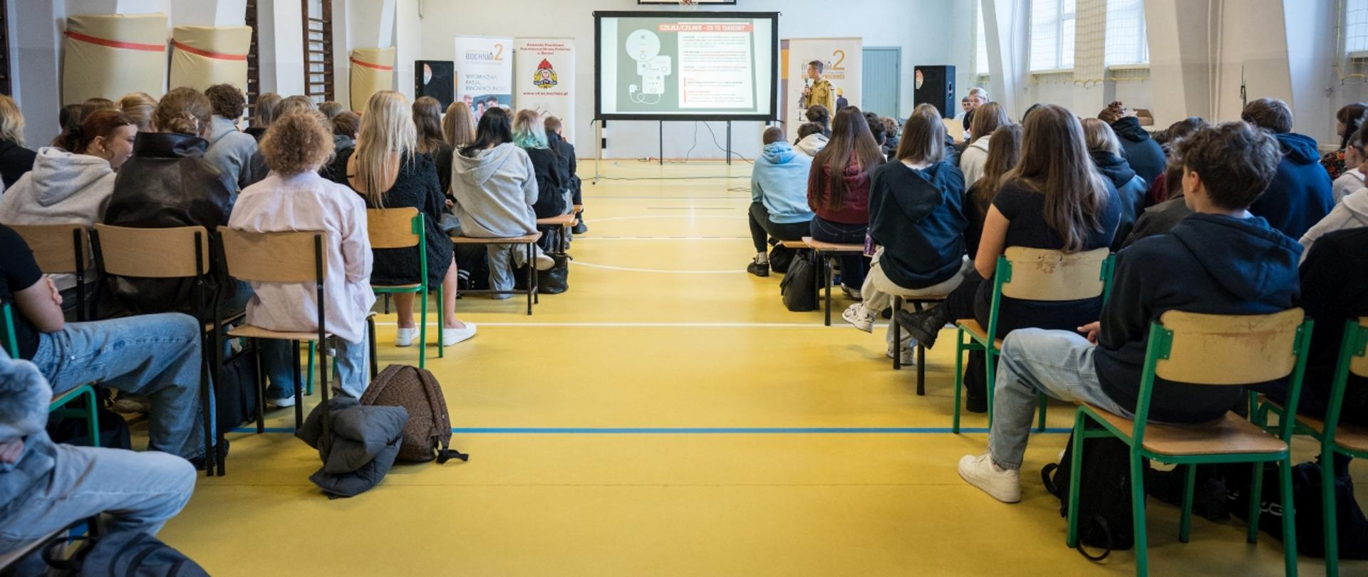 Szeroki kadr z sali gimnastycznej. Młodzież szkolna siedzi tyłem do aparatu na krzesłach i ławkach gimnastycznych, uczestnicząc w spotkaniu profilaktycznym. Na środku sali ustawiony jest duży ekran projekcyjny z prezentacją oraz prelegenci.