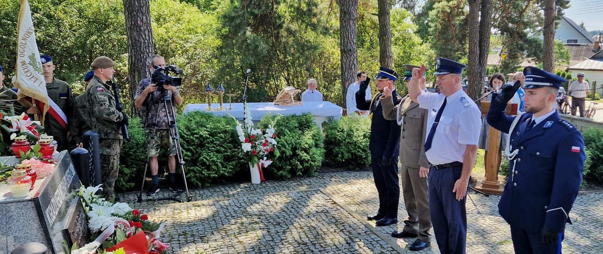 Między drzewami przed pomnikiem stoi czterech mężczyzn w mundurach - salutują.