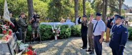 Między drzewami przed pomnikiem stoi czterech mężczyzn w mundurach - salutują.