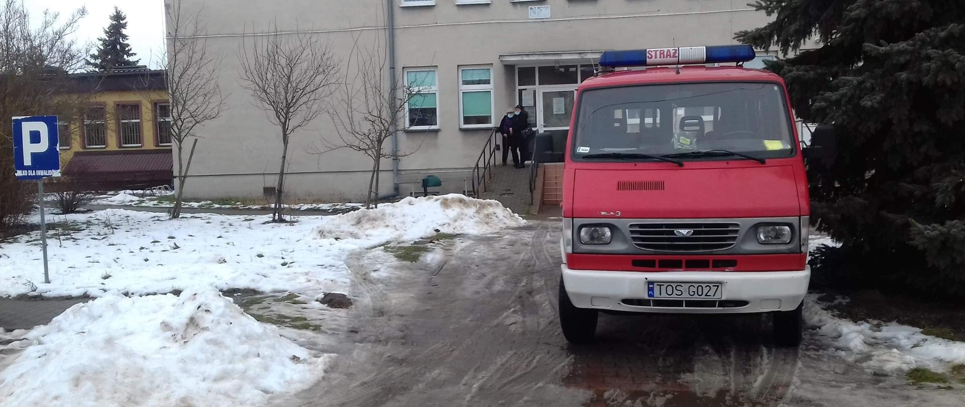Na pierwszym palnie stoi samochód pożarniczy przed przychodnią, który dowiózł pacjenta na sczepienie. Przed przychodnią stoją pacjenci. Obok samochodu rośnie świerk. Z lewej strony widać znak informujący o parkingu. 