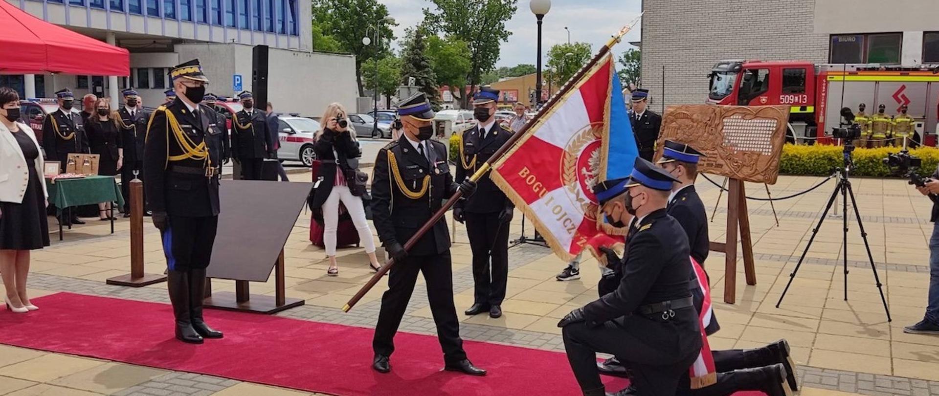 Zastępca Komendanta Głównego PSP stoi na baczność przed sztandarem , za nim stoją inni uczestnicy uroczystości. Trzymający sztandar nachyla go do przodu, przed sztandarem klęczą 3 funckcjonariusze PSP