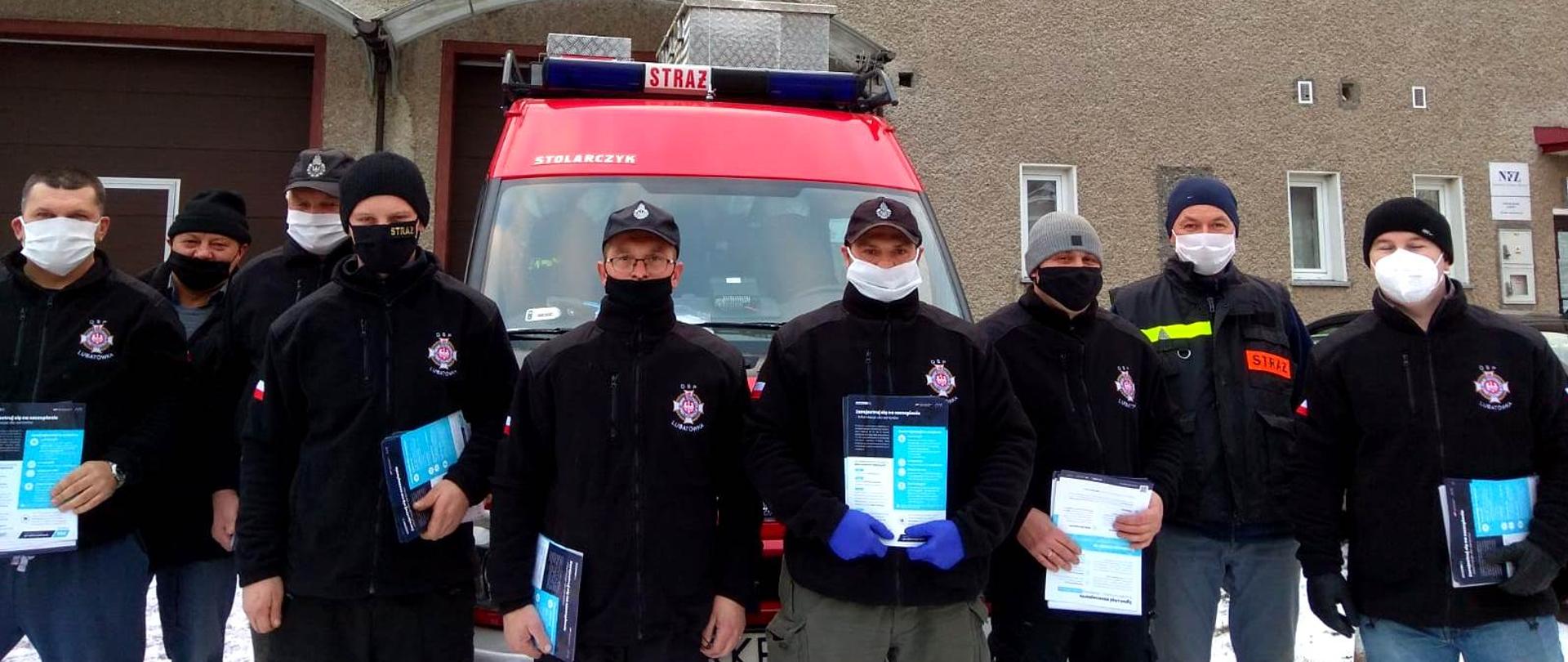 Na zdjęciu widać strażaków osm w maseczkach ochronnych stojących przed budynkiem remizy. Strażacy przygotowują się do rozwożenia ulotek informacyjnych na temat szczepień przeciw sars cov 2. Za strażakami ustawiony jest samochód pożarniczy