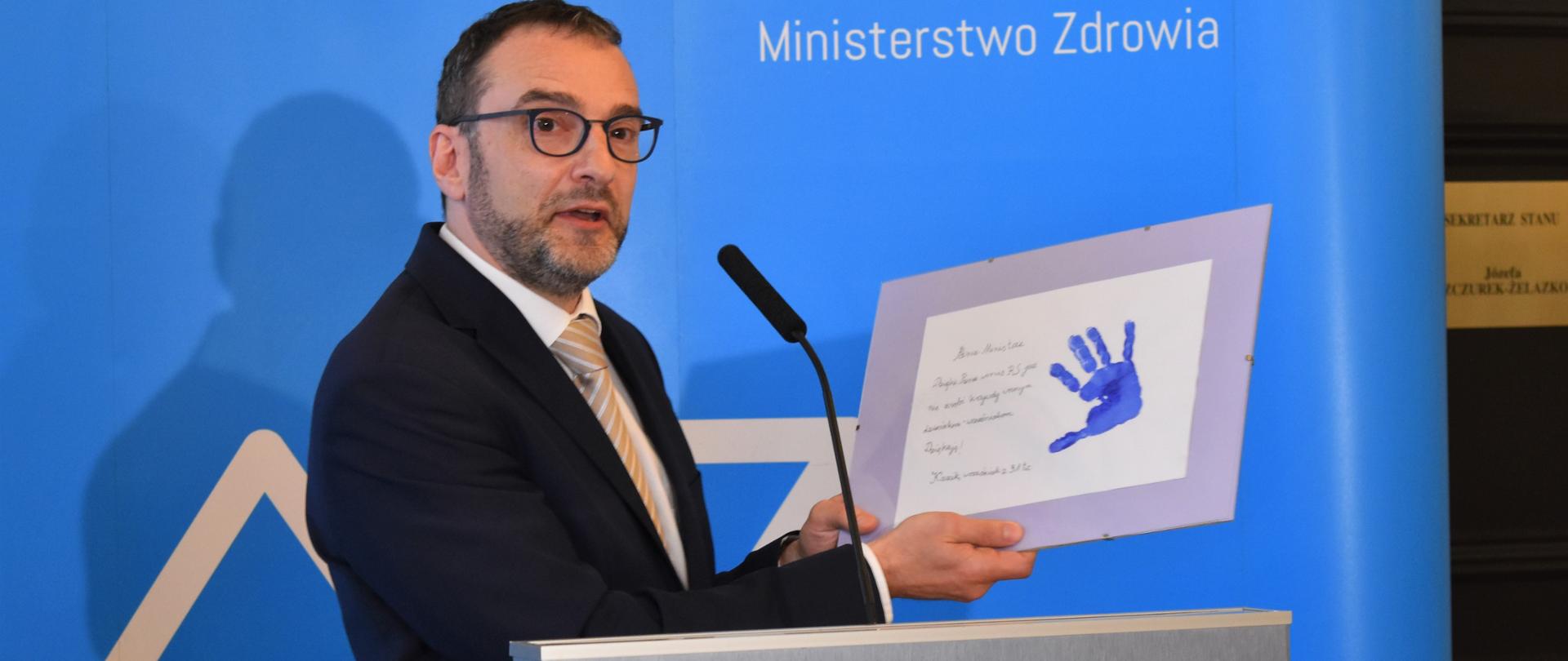 Wiceminister Marcin Czech podczas konferencji prasowej dotyczącej nowej listy leków refundowanych pokazuje podziękowania od jednego z pacjentów.