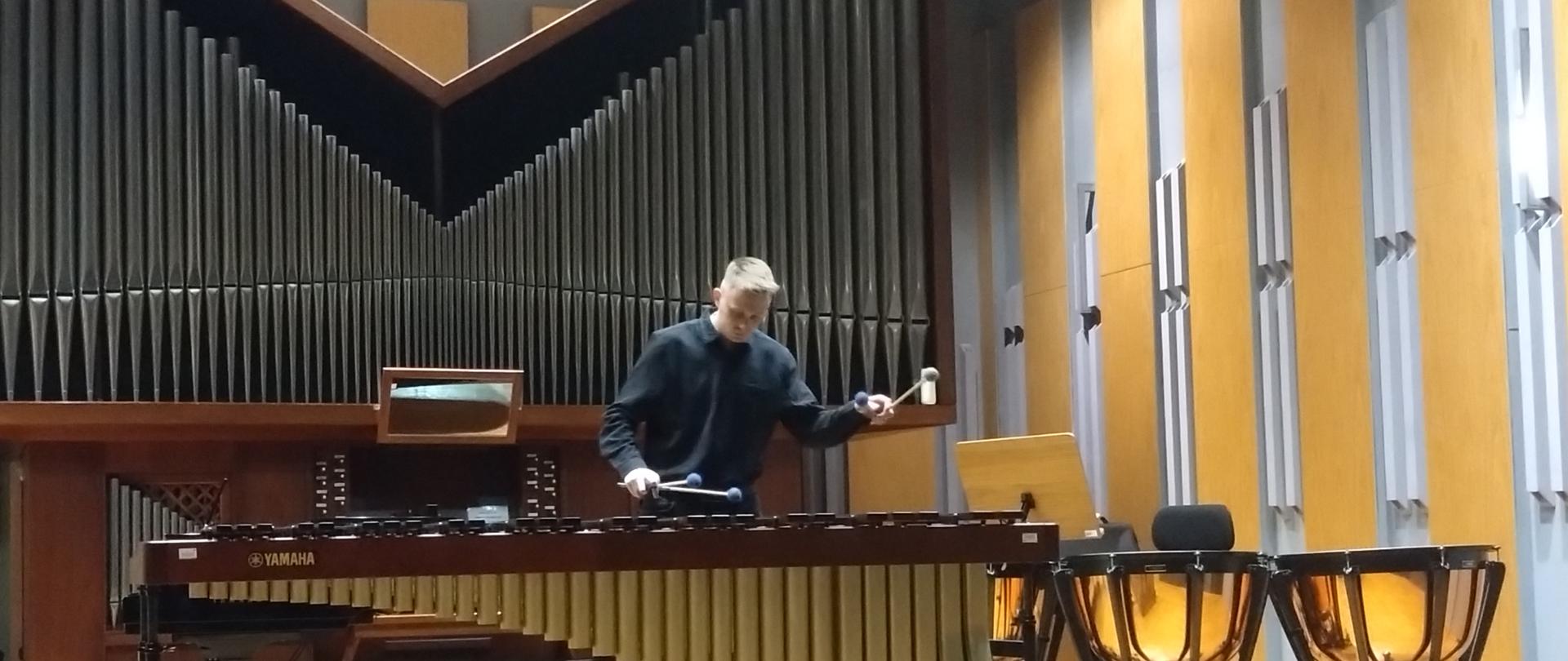 Zdjęcie w orientacji poziomej przedstawia młodego mężczyznę w trakcie występu na scenie, grającego na instrumentach perkusyjnych.
W centrum kadru stoi muzyk. Jest ubrany w czarny, długi sweter lub koszulę i czarne spodnie. Patrzy w dół, skupiony na instrumencie, trzymając w obu rękach pałki.
Gra na dużym instrumencie perkusyjnym, który zajmuje większość pierwszego planu – to marimba (lub ksylofon/wibrafon). Instrument ma dużą, ciemną ramę (widoczne logo Yamaha) i charakterystyczne, długie, drewniane rezonatory w jasnym, beżowo-brązowym kolorze, ułożone w rzędy pod klawiszami. Marimba stoi na statywie na kółkach.
Po prawej stronie, obok marimby, widoczne są kotły (timpani). Są to duże instrumenty perkusyjne z błyszczącymi, miedzianymi lub brązowymi korpusami. Są zamontowane na statywach na kółkach, a jeden z nich ma widoczną czarną naciąg.
Tło:
Scena jest oświetlona ciepłym, żółtawym światłem. Tło składa się z dwóch głównych elementów:
Po lewej stronie: Widoczny jest prospekt organowy (część piszczałkowa organów) z rzędami pionowych, ciemnych paneli. Poniżej znajduje się niższa, drewniana konsola.
Po prawej stronie: Ściana za instrumentami jest wykonana z dużych, pionowych, prostokątnych paneli w kolorze jasnego, żółtawo-pomarańczowego drewna, oddzielonych ciemniejszymi pionowymi belkami.
Muzyk stoi na jasnej, drewnianej podłodze sceny, otoczony dużymi instrumentami perkusyjnymi, przed imponującym tłem architektonicznym.