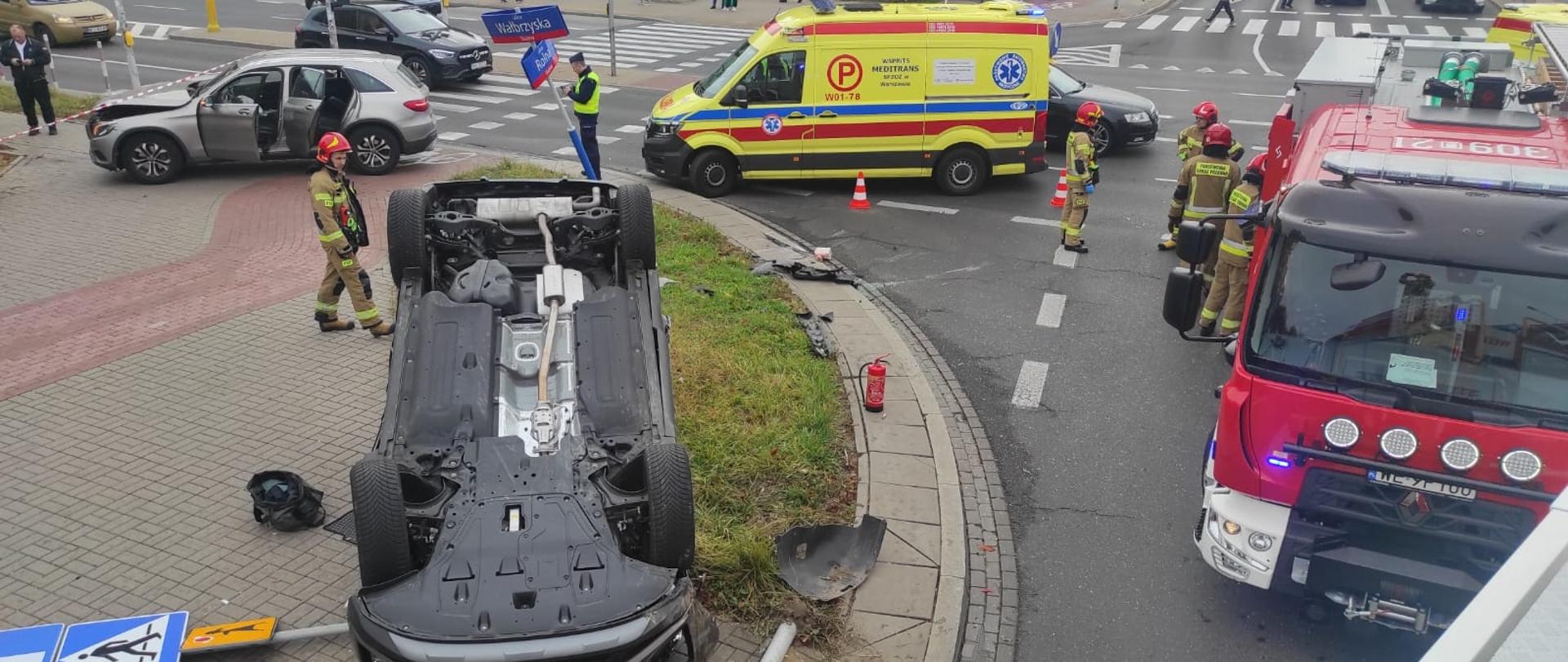 Na zdjęciu widoczne są uszkodzone samochody biorące udział w zdarzeniu. Jeden z pojazdów znajduję się na dachu. Na zdjęciu widać również pracujących strażaków oraz samochody straży pożarnej. Na zdjęciu widać również karetkę pogotowia. 