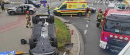 Na zdjęciu widoczne są uszkodzone samochody biorące udział w zdarzeniu. Jeden z pojazdów znajduję się na dachu. Na zdjęciu widać również pracujących strażaków oraz samochody straży pożarnej. Na zdjęciu widać również karetkę pogotowia. 